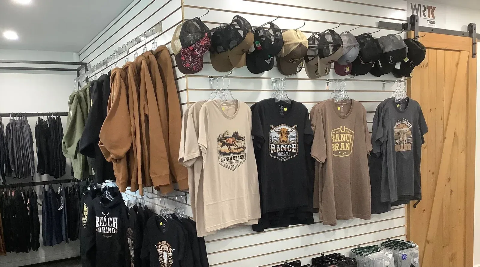 Rangée de casquettes au-dessus de t-shirts imprimés, devant une porte de grange coulissante en bois.