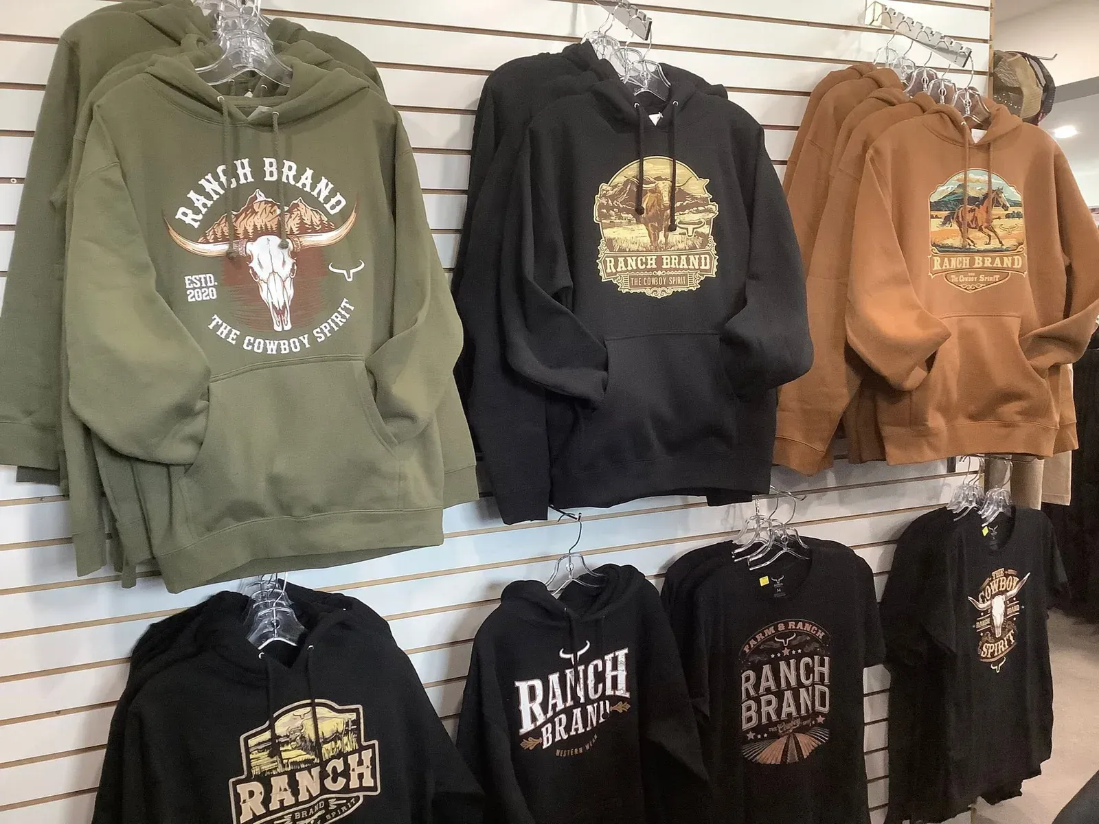 Mur à rainures garni de chandails à capuchon et de t-shirts Ranch Brand, en vert, noir et brun.