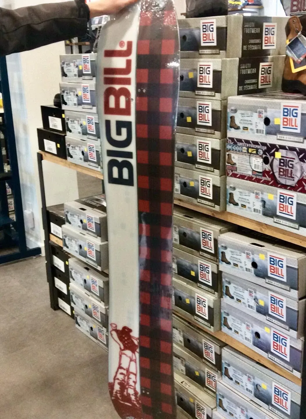 Planche à roulettes Big Bill à motif carreauté rouge et noir, appuyée contre une colonne de boîtes de bottes.